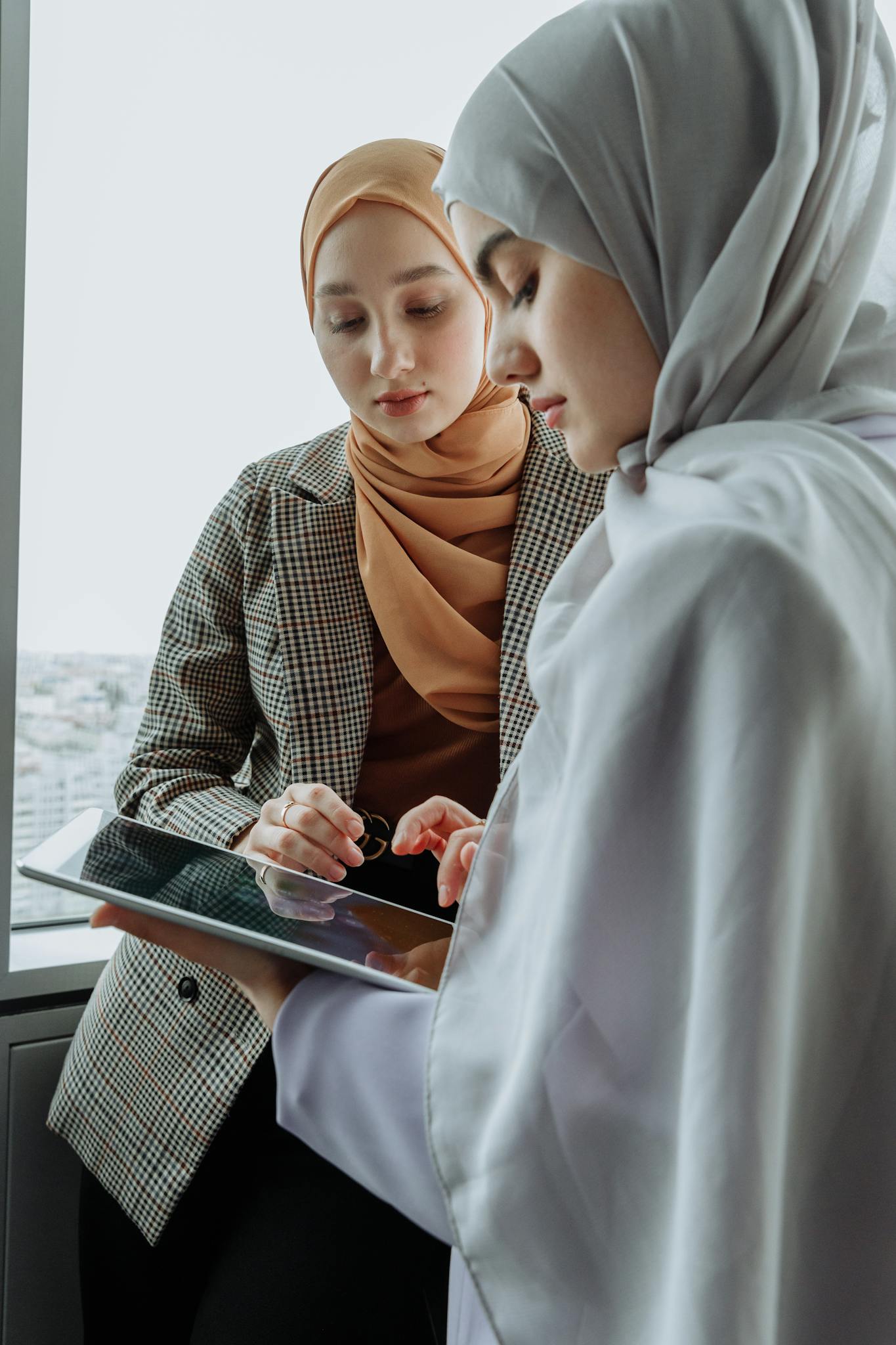 Two women in hijabs working together on a tablet in a modern office setting.